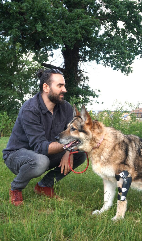 Aldi, squatting down next to a dog with a prosthetic attachment to its front left leg.