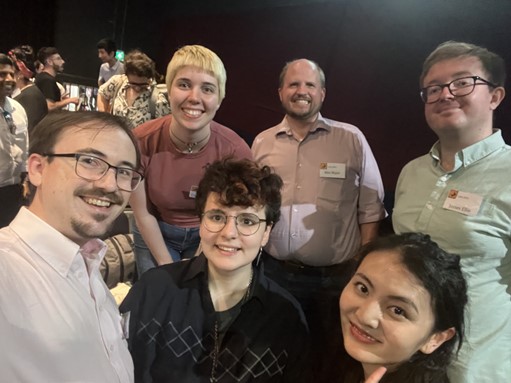 A selfie of a group of six smiling people with conference networking going on in the background.