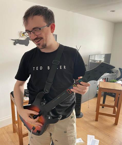A man wearing glasses and a black Ted Baker T-shirt, playing a plastic guitar-shaped video game controller.