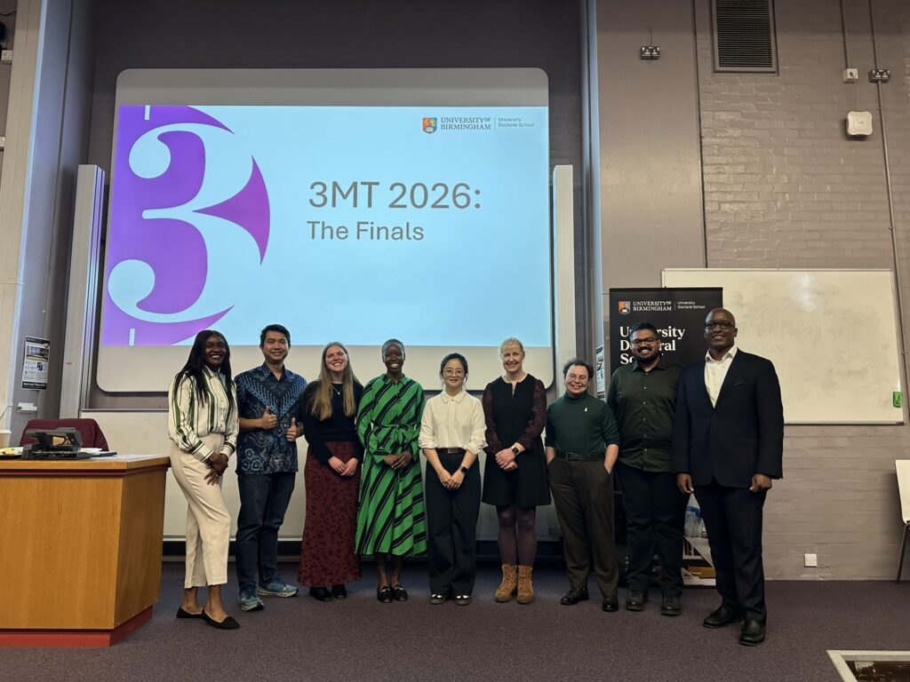 9 3MT finalists standing in a line in front of a projected slide saying "3MT 2026: The Finals".